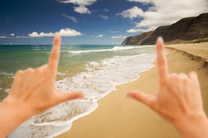 Hands framing the view of Polihale Beach on Kauaʻi, with turquoise ocean water, golden sand, and rugged cliffs in the distance under a blue sky